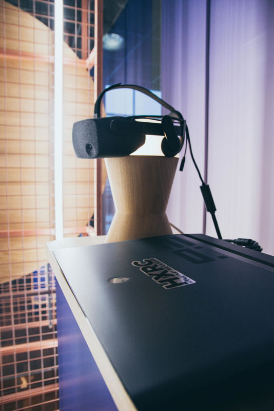 A VR headset on a wooden stand, with a laptop in the foreground. Opens in lightbox.