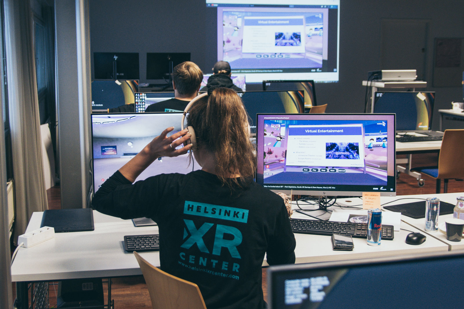 Computer room with several screens and people. In the foreground, a person is holding headphones and monitoring two screens.