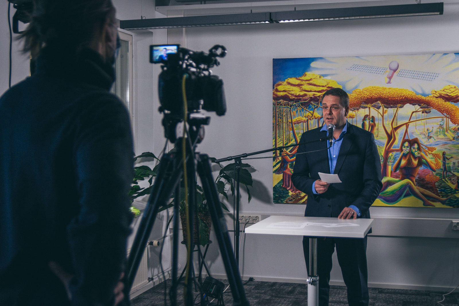 On the left there is a cameraperson with a camera on a tripod. On the right, Jan Vapaavuori, the Mayor of Helsinki, is standing in front of a microphone with a paper in his hand, speaking into the camera. Opens in lightbox.