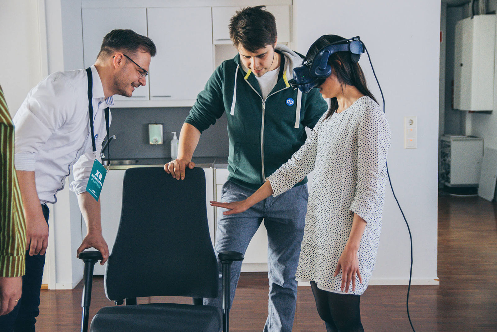 A woman trying out a virtual reality headset and reaching out. Two other people are helping her to find a chair to sit on.