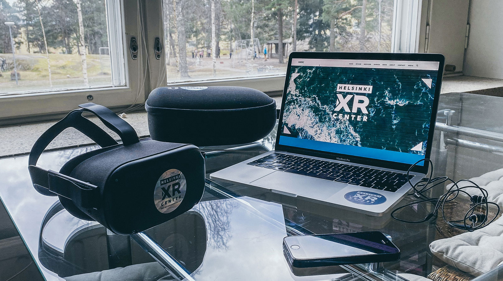 A virtual reality headset, a laptop and a phone on a table in front of a window with a view to a playground surrounded with trees.