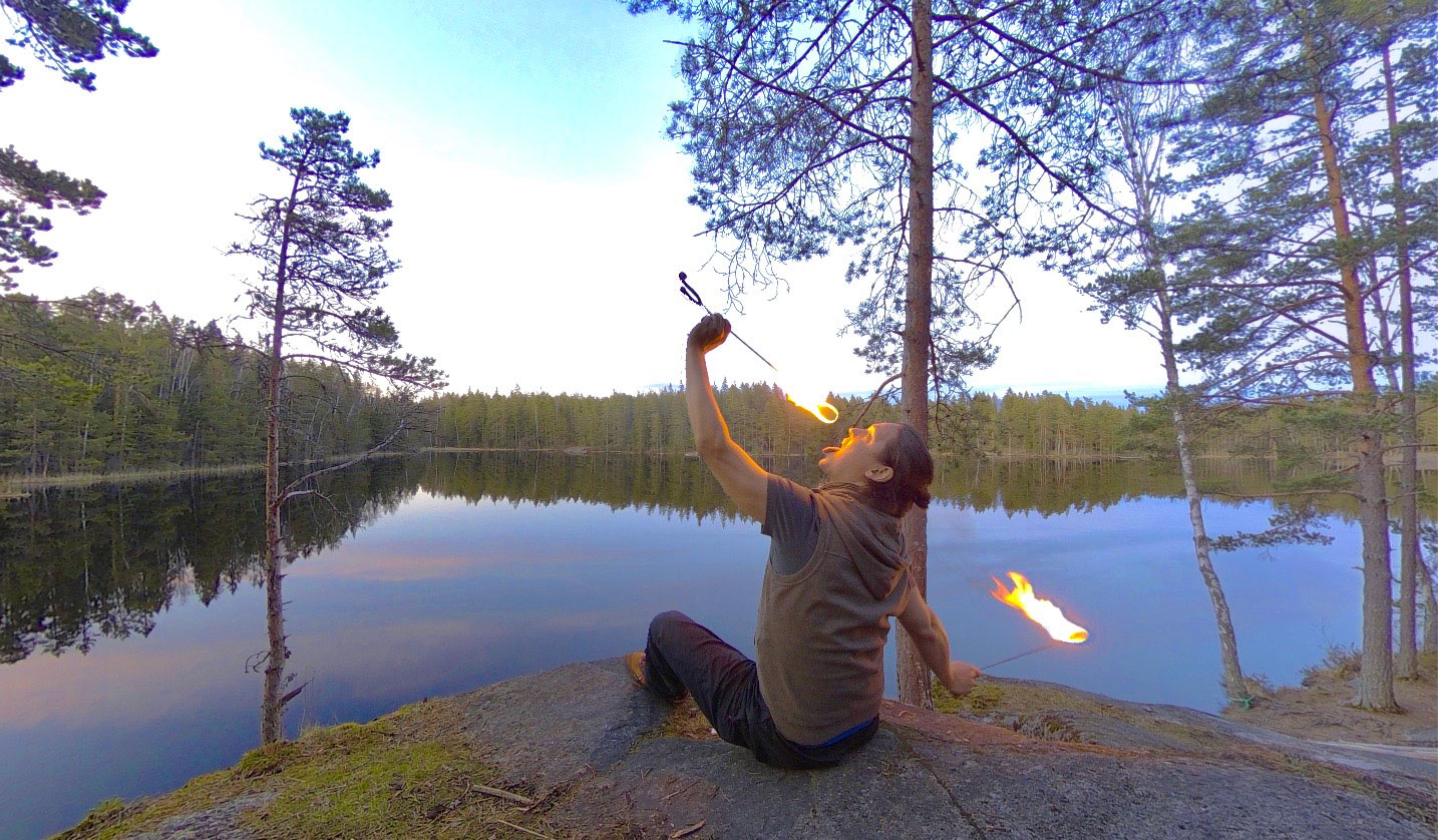Petteri Jakobsson eating fire sitting on a rock. Behind him is a very Finnish lake landscape.