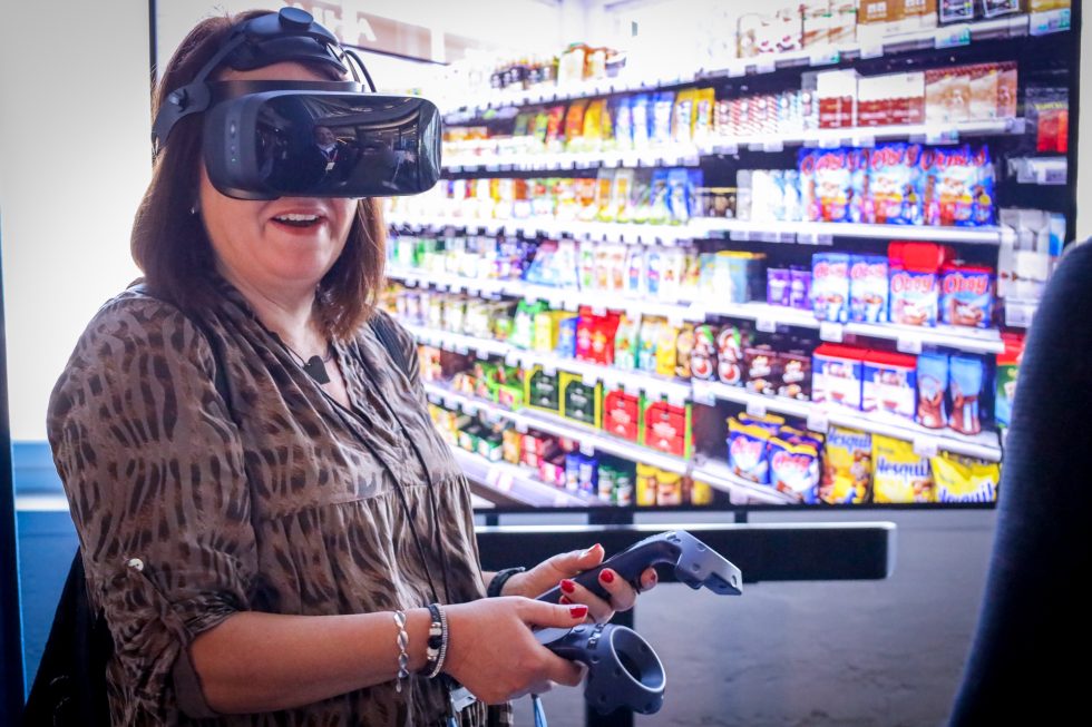 A person wearing a VR headset. The screen behind shows virtual shelving with grocery items.