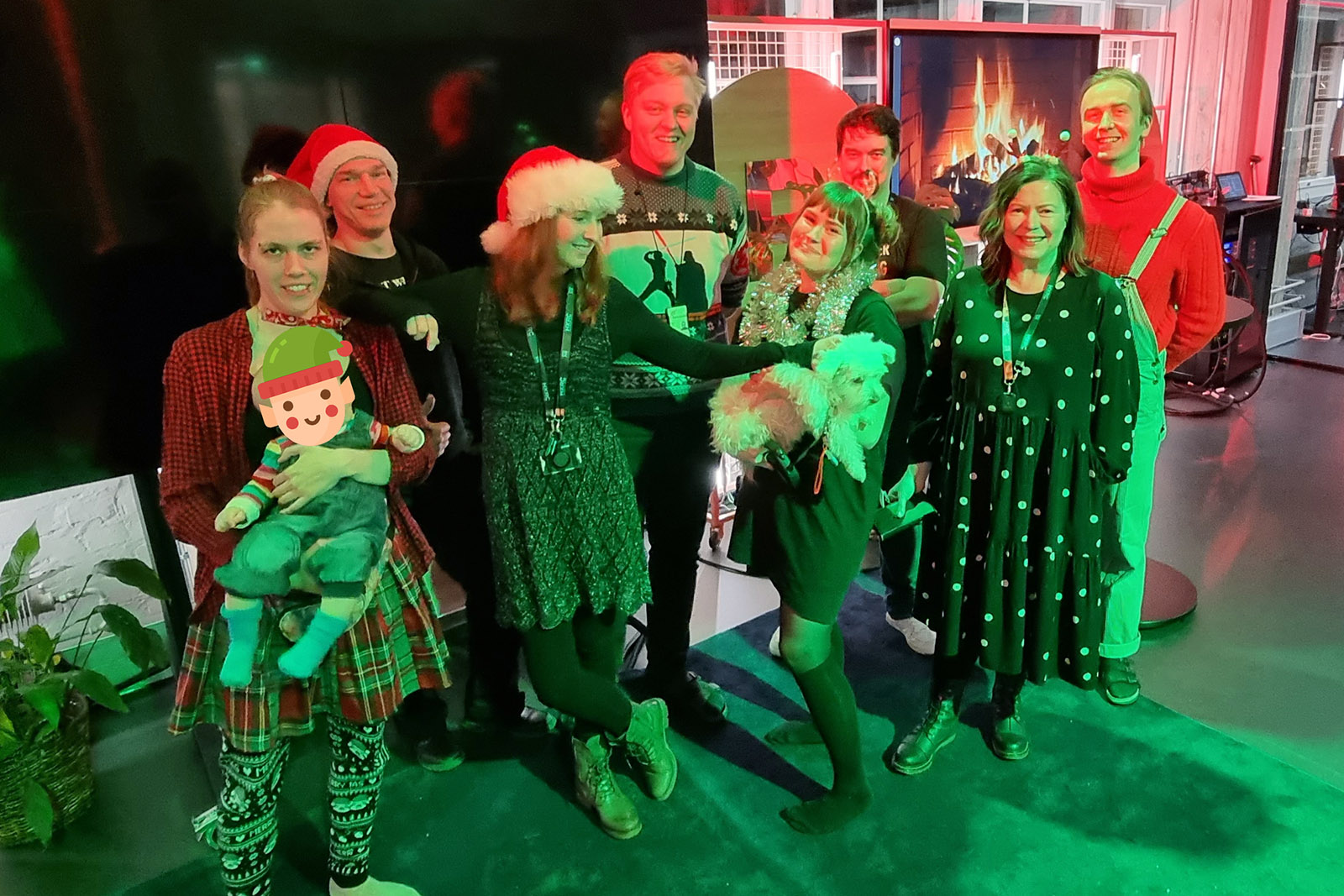 Helsinki XR Center team smiling together. One is holding a baby, and another one a small white dog. One is scratching the dog. The lighting is very christmasy: red and green.