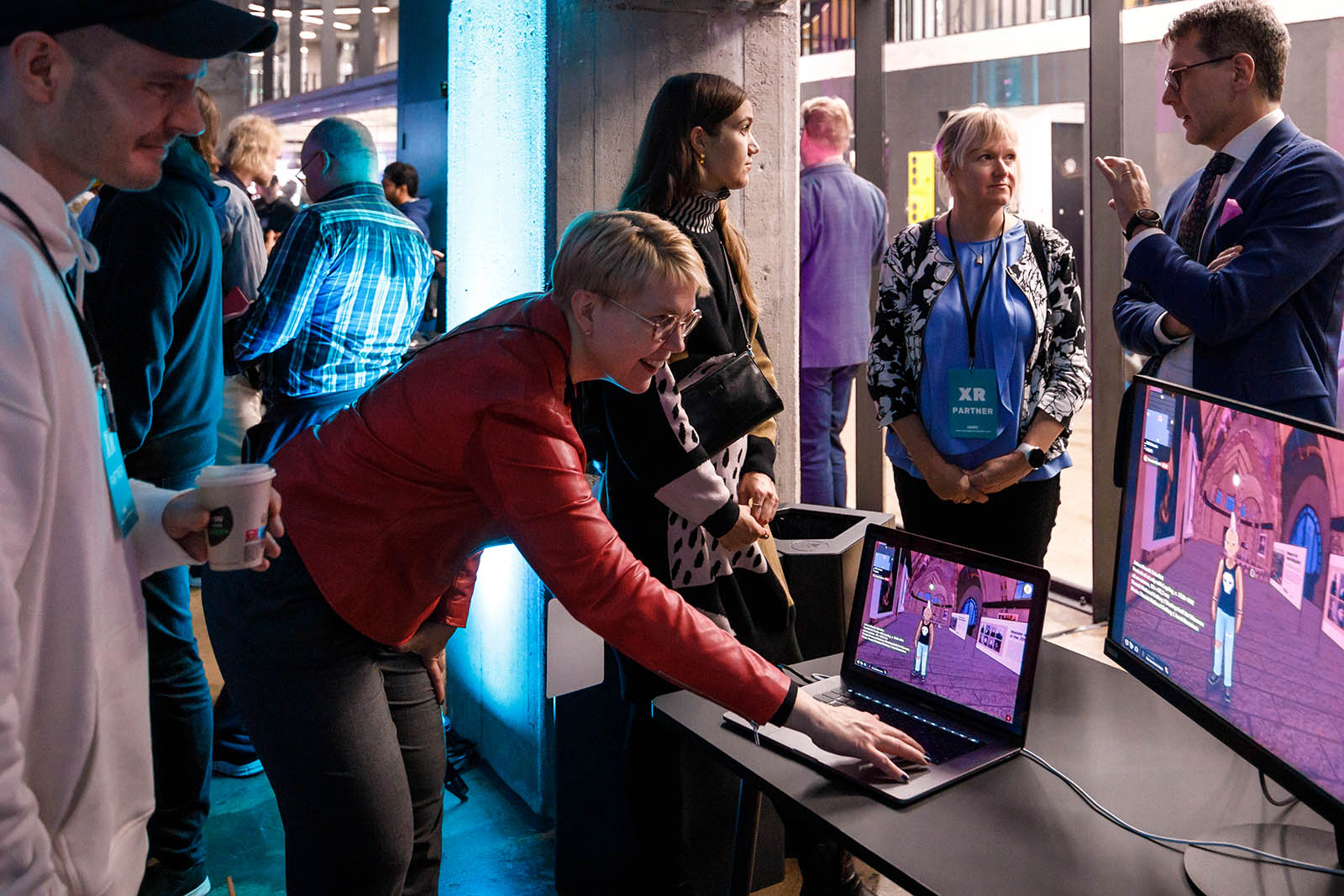 A person bent over on a laptop with a smile on their face. Another person is looking at the screen with a happy face. Other people are located in the background.