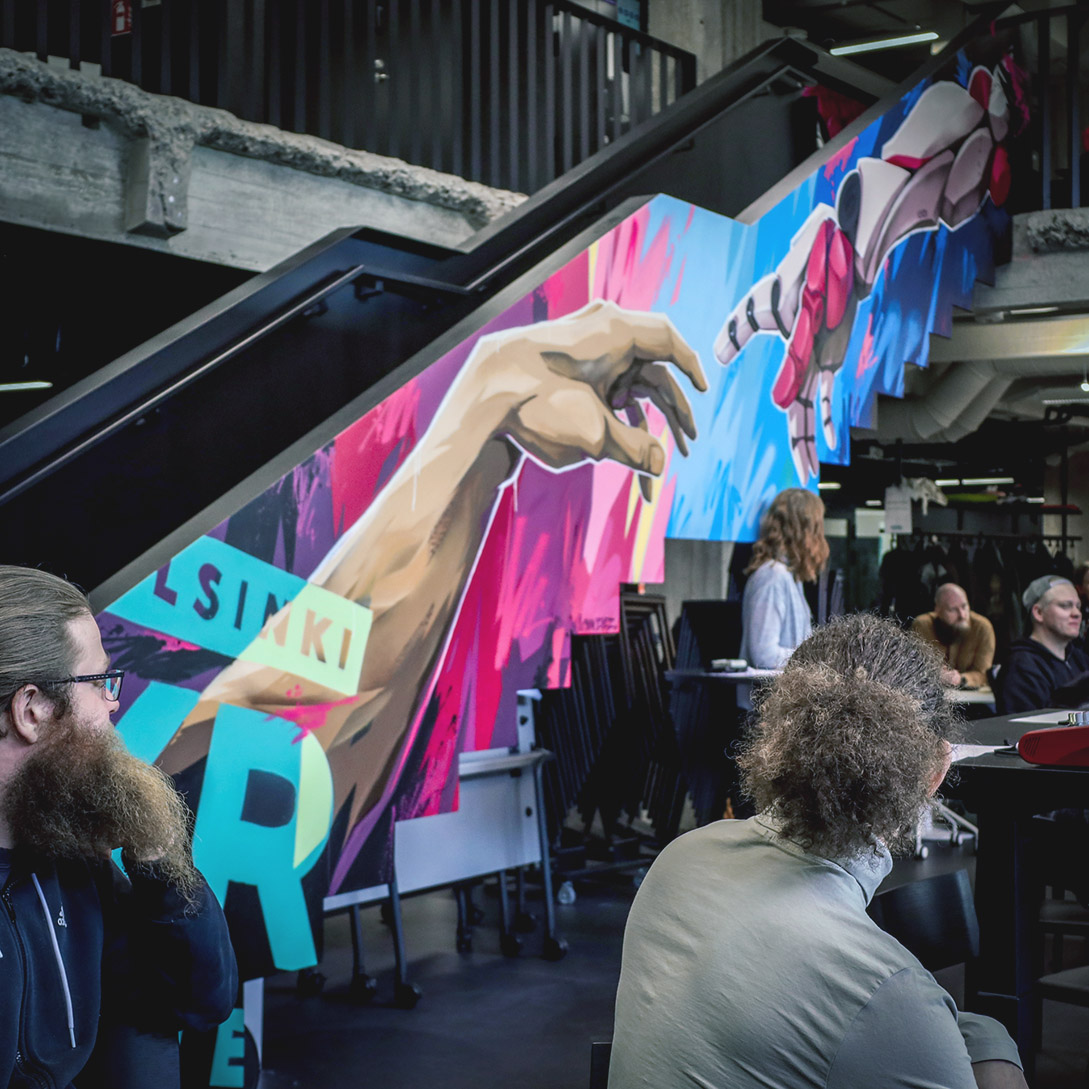 People sitting in front of a colourful graffiti located in Helsinki XR Center's common space. The graffiti is done by Mike Leggat aka Cyan Eyez during the summer of 2022.